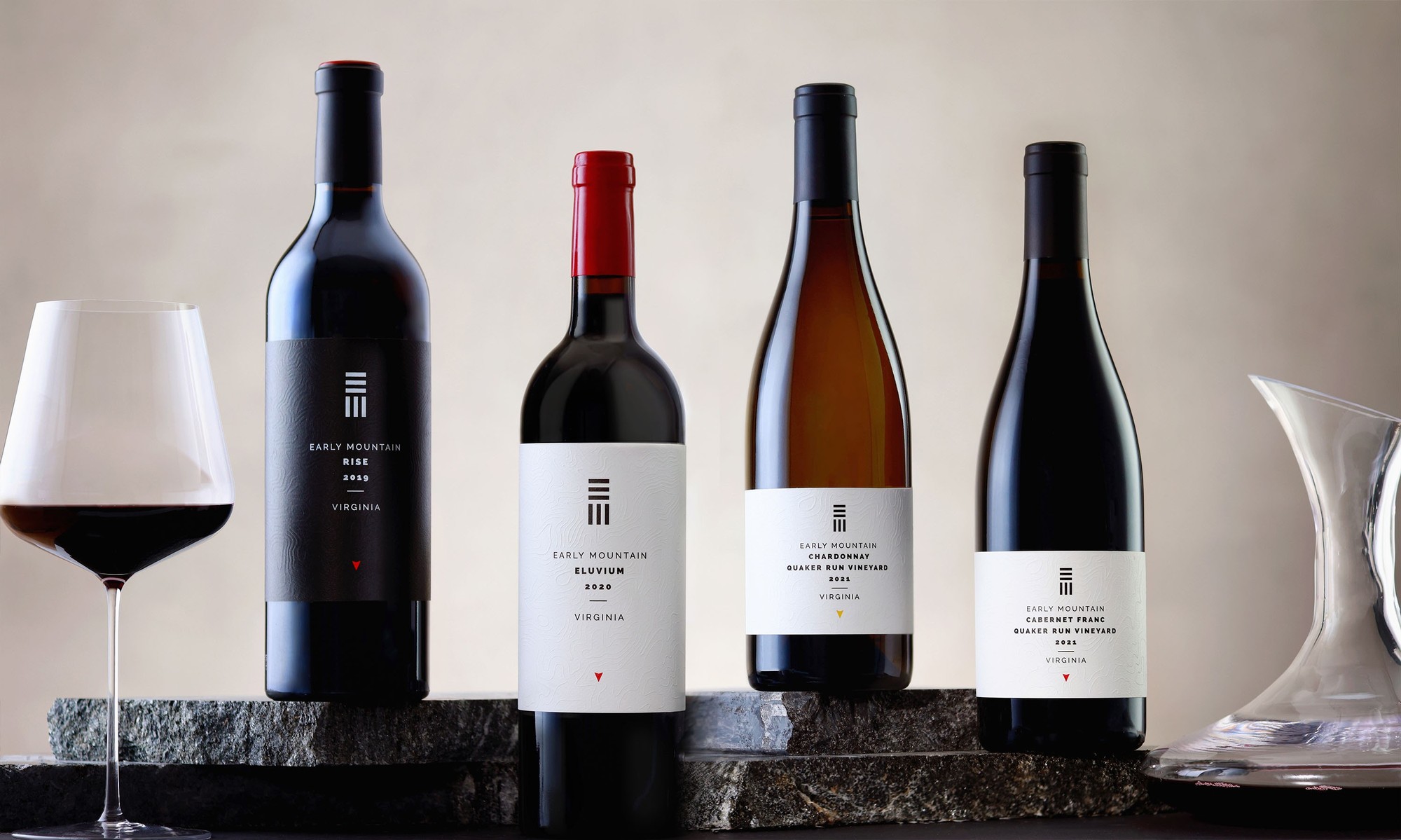Four bottles of Early Mountain Premier Wines on a dark marble slab with a filled glass on the left and a wine decanter on the right.